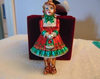 Può includere: Un addobbo natalizio in vetro a forma di figura che indossa un abito rosso con finiture verdi, una sciarpa verde e stivali marroni. La figura tiene in mano una piccola borsa argentata. L'abito presenta dettagli con glitter dorati.