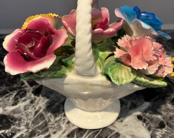 May include: A white ceramic basket with a twisted handle, filled with a sculpted floral arrangement. The bouquet features deep magenta, light pink, yellow, and blue ceramic flowers with green leaves, all set on a pedestal base.