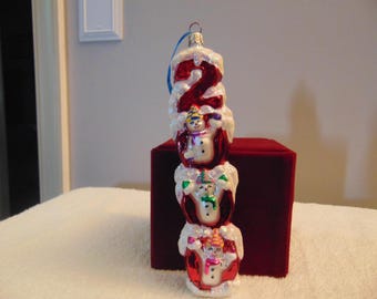 May include: A vertical glass Christmas ornament featuring the number 2 in shiny red, topped with white glittery snow. Three small snowmen with colorful hats and scarves are stacked within the red structure, each accented with white glittery snow.