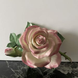 Könnte beinhalten: Eine Porzellanrosenfigur mit einer großen, offenen Blüte, deren cremefarbene Blütenblätter einen zartrosa Rand aufweisen. Die Skulptur umfasst eine kleinere, geschlossene Rosenknospe auf der linken Seite sowie detaillierte grüne Blätter.