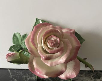 Può includere: Una statuetta di rosa in porcellana caratterizzata da un grande fiore aperto con petali color crema bordati di rosa tenue. La scultura include un bocciolo di rosa più piccolo e chiuso sulla sinistra e foglie verdi dettagliate con venature.