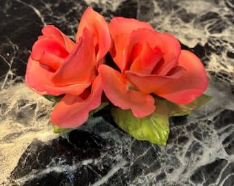 May include: A decorative ceramic figurine featuring two vibrant coral-pink roses with layered, delicate petals. The flowers are attached to a base of textured, realistic green leaves, showcasing a smooth, matte finish on the floral elements.