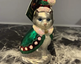 May include: A glass cat ornament featuring a silver glitter finish, wearing a green cape with red and white glitter trim, and a matching pointed hat with a white pom-pom. The cat has blue eyes and a pink nose, with a gold-toned metal cap at the top.