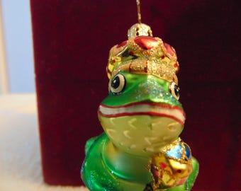May include: A glass frog ornament featuring a bright green body, a wide mouth with a white and red line, and large black eyes. The frog wears a gold crown with red accents and glitter, and holds a small, colorful, glittery object in one hand.
