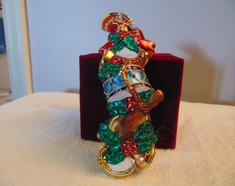 May include: A glass Christmas ornament shaped like a stack of drums and a trumpet, decorated with green holly leaves, red berries, and gold accents. The ornament features a shiny finish with blue and brown details and a metallic gold hanging loop.
