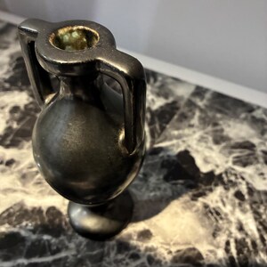 Pu&ograve; includere: Un piccolo vaso in ceramica nera in stile anfora con due manici verticali che si estendono dal collo alla spalla. Il vaso presenta un corpo arrotondato, un collo stretto e una base circolare. L'interno dell'apertura mostra una smaltatura verde maculata.