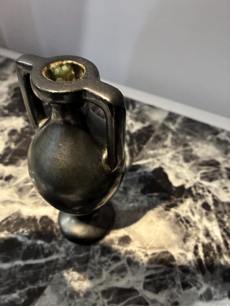 Pu&ograve; includere: Un piccolo vaso in ceramica nera in stile anfora con due manici verticali che si estendono dal collo alla spalla. Il vaso presenta un corpo arrotondato, un collo stretto e una base circolare. L'interno dell'apertura mostra una smaltatura verde maculata.