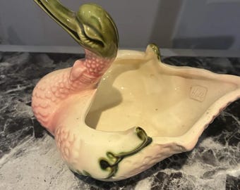 May include: A ceramic swan-shaped planter featuring a glossy finish with a pale pink body and a green head and neck. The interior is cream-colored and includes a stamped Hull mark on the inner wall. The exterior surface has a textured, dimpled pattern.