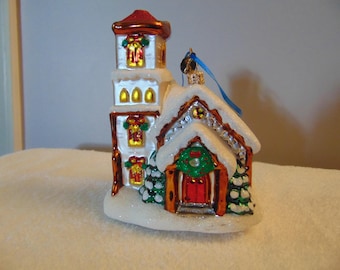 May include: A glass Christmas ornament shaped like a small church with a tall tower, red doors, and a green wreath. The structure features white glittery snow on the roof and base, with gold-trimmed windows and small green pine trees.