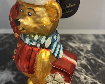 May include: A glass Christmas ornament shaped like a golden bear wearing a red sweater, a blue and white striped scarf, and a matching hat. The bear holds a red gift box with a white bow. A black circular tag with the text Christopher Radko is attached.