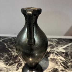 Pu&ograve; includere: Un piccolo vaso in metallo scuro con una finitura liscia e riflettente. Il recipiente presenta un collo stretto, un unico manico verticale che si estende dal bordo alla spalla e un corpo arrotondato che si restringe verso una base.