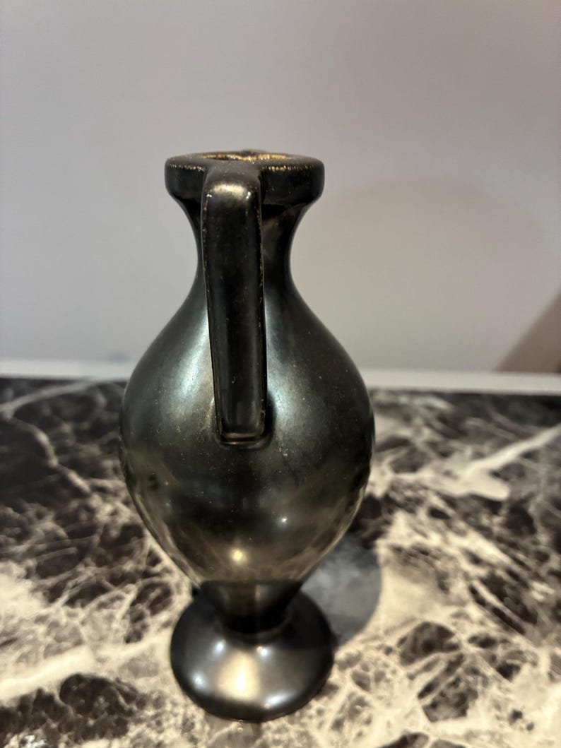 Pu&ograve; includere: Un piccolo vaso in metallo scuro con una finitura liscia e riflettente. Il recipiente presenta un collo stretto, un unico manico verticale che si estende dal bordo alla spalla e un corpo arrotondato che si restringe verso una base.