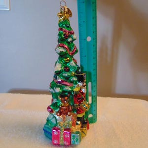 Könnte beinhalten: Ein Weihnachtsbaumschmuck aus Glas in Form eines grünen Baumes mit roten und goldenen Akzenten, einem Nussknacker, einem Teddybären und Geschenken am Fuß. Ein grünes Lineal daneben zeigt eine Höhe von 18 cm an.