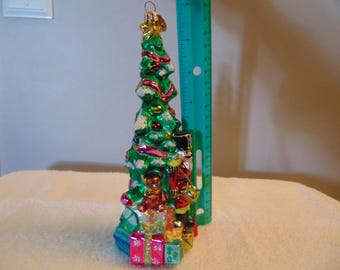 May include: A glass Christmas tree ornament featuring a green tree decorated with red and gold accents, a nutcracker, a teddy bear, and wrapped gifts at the base. A green ruler is placed next to the ornament, showing a height of 7 inches.