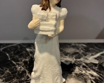 May include: A porcelain figurine of a person with brown hair wearing a long white dress with puffed sleeves. The figure is holding a stack of white rectangular blocks in their arms. The sculpture has a glossy finish and stands upright.