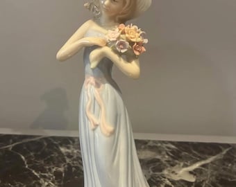 May include: A porcelain figurine of a person wearing a wide-brimmed hat and a long, pale blue dress with a pink sash. The figure holds a bouquet of small, multi-colored flowers in their hands, with a glossy finish and delicate facial features.