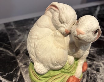 May include: A ceramic figurine featuring two white rabbits huddled together. One rabbit is resting with its eyes closed, while the other is positioned beside it. They are perched on a base painted with green textured grass and orange accents.