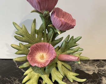 Può includere: Una statuetta decorativa in ceramica raffigurante tre fiori color malva con petali testurizzati e centri gialli, circondati da dettagliate foglie verdi seghettate e un singolo bocciolo chiuso, disposti in un grappolo botanico verticale.