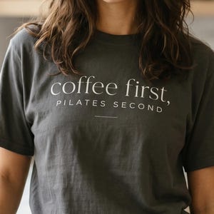 May include: A charcoal grey short-sleeved t-shirt featuring the white text "coffee first, PILATES SECOND" printed on the chest. The shirt has a crew neckline and a relaxed fit, paired with solid black athletic shorts.