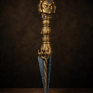 Puede incluir: Una daga ritual con un mango de color dorado que presenta un rostro esculpido, calaveras y detalles de piedras azules. La hoja es de color gris oscuro con una textura similar a la piedra y una serpiente dorada enroscada en la base.