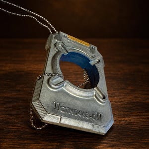 Op de afbeelding: Een zilverkleurige rechthoekige hanger met een centrale ronde uitsparing en een blauwe binnenrand. Het stuk heeft een metallic afwerking, een klein goudkleurig accent aan de bovenkant en de in reliëf aangebrachte tekst IICTN0463-0II. Het hangt aan een zilveren bolletjesketting.