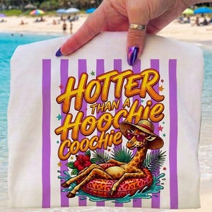 Può includere: Telo mare bianco con strisce verticali viola e la scritta "Hotter Than a Hoochie Coochie". Il telo presenta una giraffa con occhiali da sole e cappello, sdraiata su un galleggiante in acqua. È visibile anche un fiore di ibisco rosso.