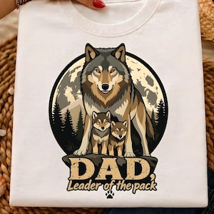 May include: White t-shirt featuring a wolf family design. The graphic includes a wolf, two cubs, and the text "DAD Leader of the pack" in a distressed font. The design is set against a moonlit background with trees.