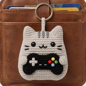 May include: Crocheted cat keychain in gray with a black video game controller. The cat has black eyes, whiskers, and gray stripes. The controller has colored buttons. Attached to a metal keyring.