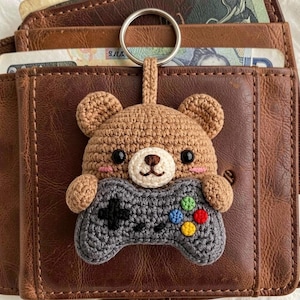 May include: A crocheted brown bear keychain holding a gray video game controller. The bear has black eyes, a white muzzle, and pink cheeks. The controller has colorful buttons. The keychain is attached to a silver ring.