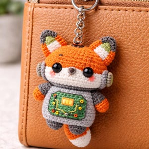 May include: Crocheted fox keychain with orange, gray, and white details. The fox has large black eyes, a circuit board design on its chest, and is attached to a silver keychain. A cute and unique accessory.