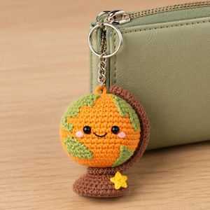May include: A crocheted globe keyring with a smiling face, green continents, and a yellow star. The orange and brown globe is attached to a silver keyring, next to a green purse.