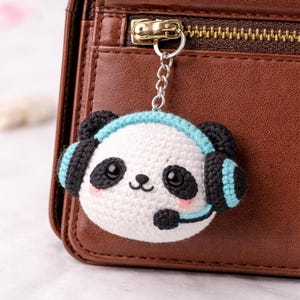 May include: Crocheted panda bear keyring with black and blue headphones. The panda has black eyes, ears, and a microphone. The keyring is attached to a brown leather wallet with a gold zip.