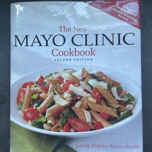 May include: The New Mayo Clinic Cookbook, second edition, featuring award-winning recipes. The cover displays a bowl of pasta with tomatoes, spinach, and beans. The book's title and author are prominently displayed.