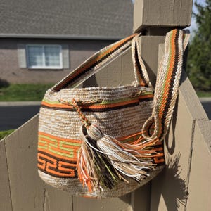 May include: Handmade crochet shoulder bag with a cylindrical shape. The bag features a beige base with horizontal stripes in orange, green, and brown. A long strap and a decorative tassel with multiple colors complete the design. The bag is hanging on a wooden post.