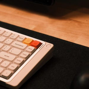 May include: A cream-colored mechanical keyboard with white keycaps and orange and red accent keys. The keyboard is on a black surface, with a black mouse and wooden desk in the background.