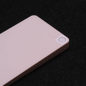 May include: A rectangular, light pink electronic device with a smooth, matte finish. The device features a single, circular white button positioned in the top right corner, set within a slightly raised square area on the flat surface.