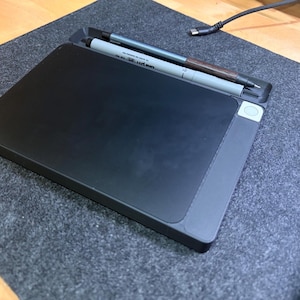 Puede incluir: Una tableta negra con un portalápices incorporado, que contiene un bolígrafo plateado y otro marrón. La tableta está sobre una superficie gris, con un cable negro y una superficie de madera en el fondo. La tableta tiene un botón lateral.