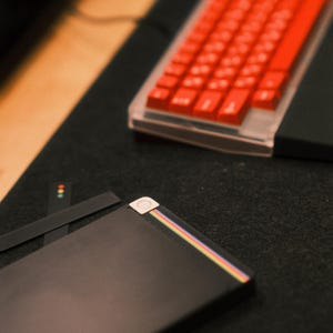 Puede incluir: Primer plano de un teclado mecánico con teclas rojas y una base transparente. Un dispositivo negro con una franja de arcoíris está en primer plano. Los elementos están sobre una superficie oscura.