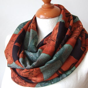May include: A patterned infinity scarf in shades of rust orange, teal, and black. The scarf is displayed on a mannequin, highlighting its circular shape and fabric. The design includes abstract shapes and lines.