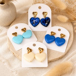 Pode incluir: Brincos em forma de coração em várias cores: azul claro, azul escuro e creme. Cada par apresenta um pino em forma de coração dourado. Os brincos são exibidos em cartões brancos.