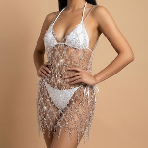 May include: White bikini set with a beaded, see-through cover-up. The bikini top has halter straps and the bottoms have tie sides. The cover-up is made of clear beads that hang down in a fringe-like pattern.