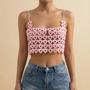 May include: Pink crop top with a heart-shaped design. The top features a square neckline and pearl-like straps. The garment is made of connected heart shapes and is worn with blue denim shorts.