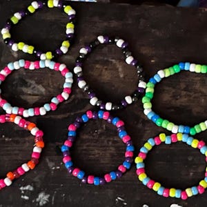 May include: A collection of six circular beaded bracelets featuring various color combinations. The bracelets are constructed from small, round, opaque plastic beads in shades of neon green, purple, black, white, pink, blue, orange, and yellow.