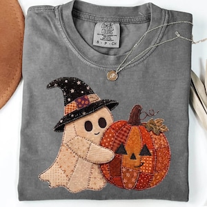 May include: A charcoal gray t-shirt featuring a stitched applique design of a light beige ghost wearing a black star-patterned witch hat, hugging a patchwork pumpkin made of various orange and red fabric patterns with a smiling jack-o'-lantern face.