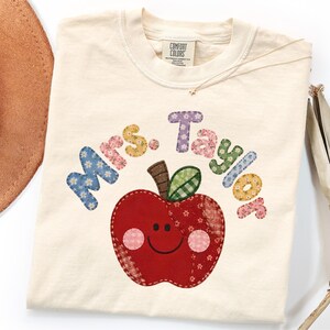 May include: An ivory t-shirt featuring a graphic of a red apple with a smiling face and pink cheeks. The apple has a brown stem and a green leaf. Above the apple, the text Mrs. Taylor is printed in a multi-coloured, floral-patterned font.