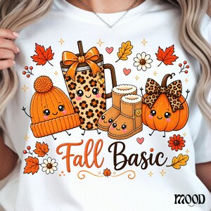 May include: A white t-shirt featuring a graphic print of a leopard print tumbler, an orange knit beanie, tan boots, and an orange pumpkin, all with smiling faces. The text Fall Basic is printed in orange and black script below the items.