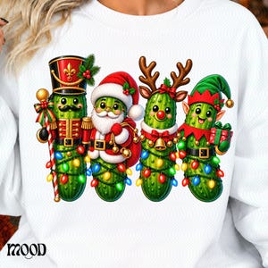 Puede incluir: Una sudadera blanca con un gráfico de cuatro pepinillos verdes vestidos como personajes navideños: un cascanueces, Santa Claus, un reno y un elfo. Cada pepinillo está envuelto en luces de colores. La palabra MOOD está impresa en negro en la parte inferior.