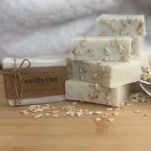 Può includere: Una pila di saponi rettangolari di colore chiaro con avena incorporata. Un fascio di sapone avvolto con un'etichetta marrone e il testo "Vanilla Oat" è legato con spago. Sono visibili anche una piccola ciotola di vetro con avena e un cucchiaio di legno.
