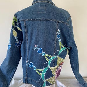 May include: A blue denim jacket with hand-painted floral and geometric designs in shades of green, blue, and purple. The jacket features a classic collar, button closure, and long sleeves. The back of the jacket is the focal point, showcasing the colorful artwork.