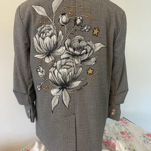 May include: A black and white houndstooth blazer featuring a large back graphic of three detailed peonies, a golden crescent moon, and small stars. The design includes a handwritten signature at the bottom right of the floral illustration.